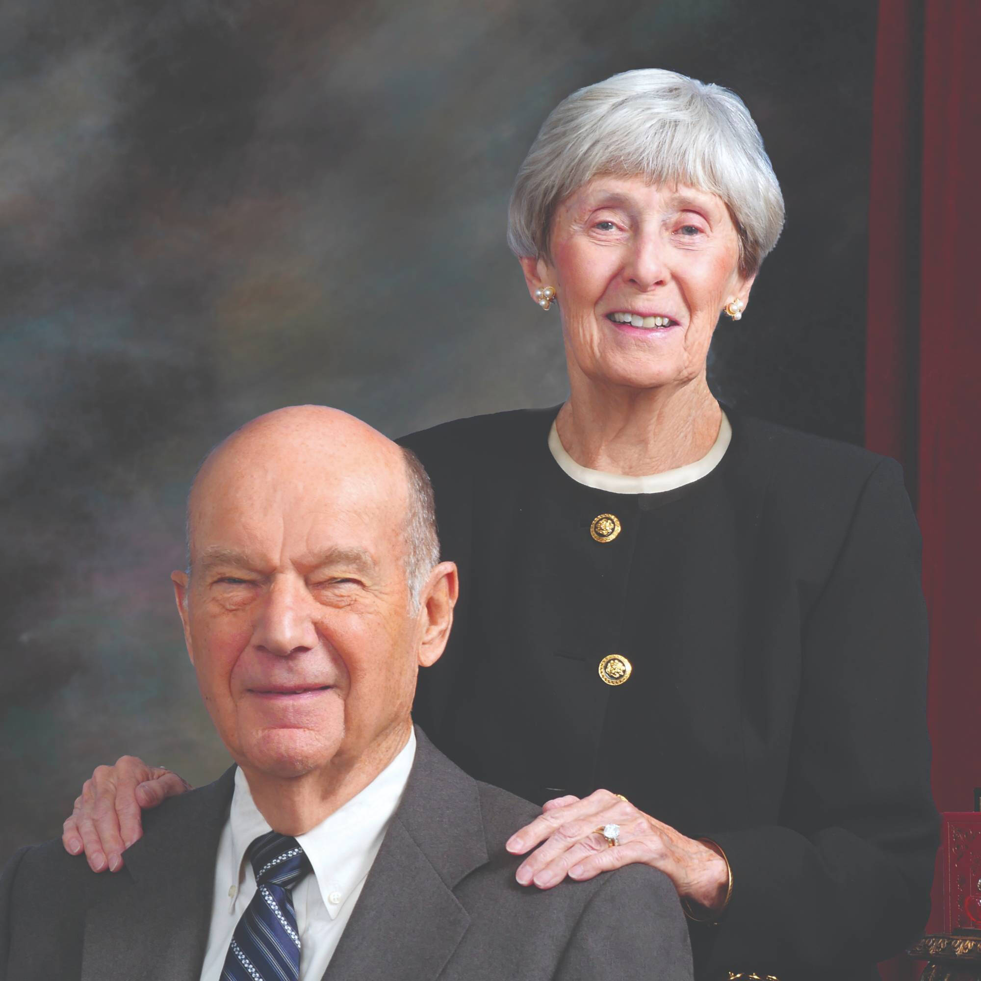 A photo of Bob Thompson seated with his wife, Ellen Thompson, standing behind him with her hands on his shoulders, both are smiling.